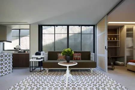 Modern minimalist living room with green sofa, white coffee table topped with potted flowers, geometric rug, and sliding glass doors to adjacent kitchen and bedroom. - Bali Villas