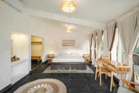 Spacious tropical bedroom with white bedding on a wooden king bed, rattan pendant lights, wooden desk and chairs, and large curtained windows.