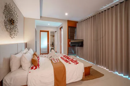 Luxurious hotel bedroom with king bed scattered in red rose petals, folded towels, wall-mounted TV, and tropical wooden accents.