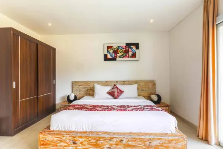 Cozy Balinese bedroom with rustic wooden bed, white linens, red throw, abstract wall art, and sliding glass doors.