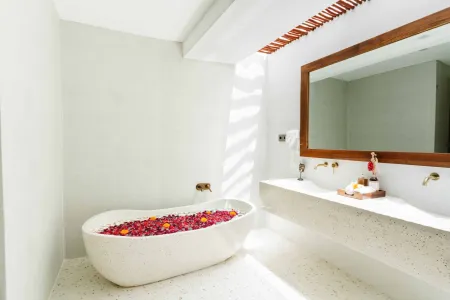 Luxurious spa bathroom with white freestanding bathtub filled with floating red hibiscus petals, wooden mirror, and brass fixtures in soft natural light. - Bali Villas