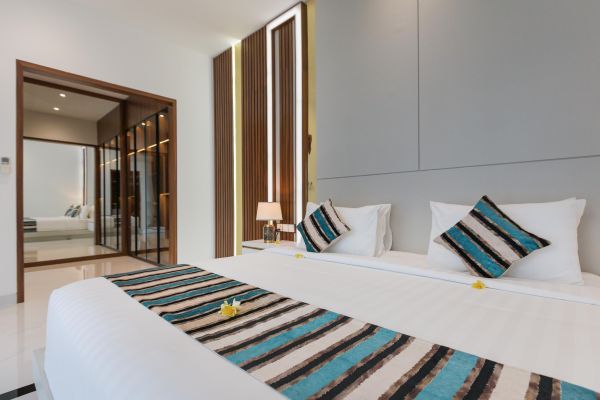 Luxurious tropical bedroom with king bed, white linens, teal striped runner, frangipani flower, and wooden doors to balcony in modern Bali villa style.