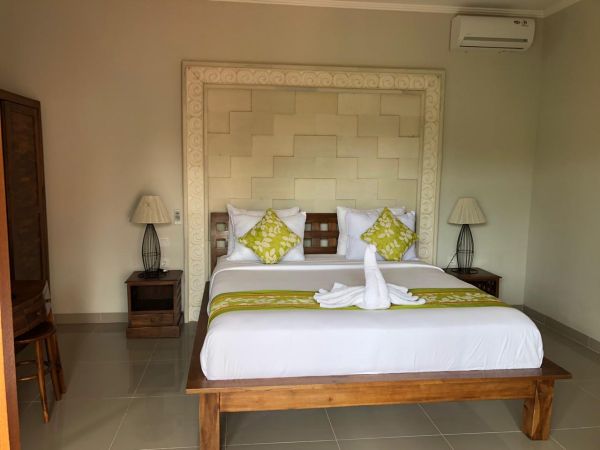 Cozy Balinese hotel bedroom with king bed, green accents, swan-folded towel, wooden headboard, and bedside lamps.