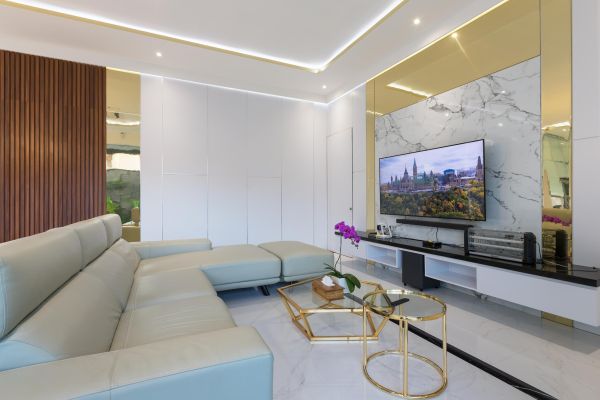 Modern living room with light gray L-shaped sofa, gold glass coffee table, large TV on white media unit against marble wall, wooden accents, and purple orchids. - Bali Villas