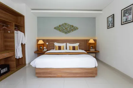 Elegant Balinese hotel bedroom with wooden king bed, mustard pillows, bedside lamps, open wardrobe with robe, and green floral wall decor. - Bali Villas