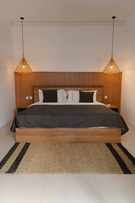 Modern bedroom with wooden bed, gray bedding, woven pendant lights, and striped rug on white floor.