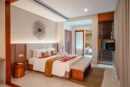 Luxurious Balinese-style bedroom with a king bed adorned in red rose petals, wooden accents, ambient lighting, and an open bathroom door.