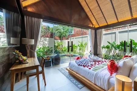 Luxurious Bali villa bedroom with rose petal heart on king bed, towel swans, wooden desk with fruits, and sliding doors to lush tropical garden.