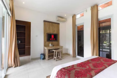 Spacious Bali villa bedroom featuring a wooden desk, Netflix TV, ornate red bedspread, and sliding doors to a garden balcony.