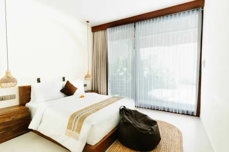 Serene Balinese bedroom with white linens on a wooden bed, woven pendant lights, sheer curtains on sliding doors to tropical garden, and a black duffel bag on a round rug.