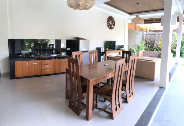 Modern open-plan kitchen and dining area with wooden table, chairs, black cabinets, and tropical garden views. - Bali Villas