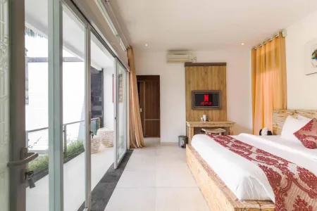 Spacious tropical bedroom with wooden bed in red bedding, wall-mounted TV, orange curtains, and sliding glass doors to balcony with plants.