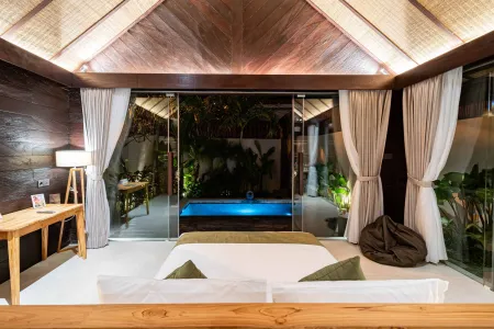 Spacious tropical villa bedroom with wooden accents, king bed, desk, and sliding doors opening to a lit infinity pool and lush garden.