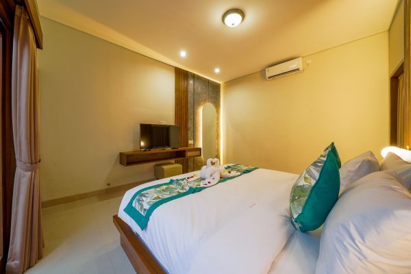 Cozy Balinese hotel bedroom with king bed, green pillows, towel swan decor, TV, and warm lighting. - Bali Villas