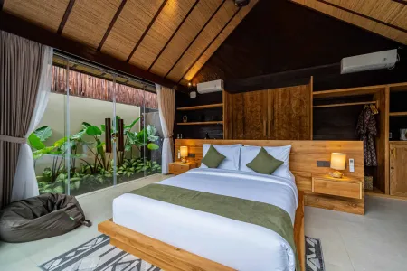 Serene Balinese bedroom with wooden platform bed, white linens, tropical plants, and large windows overlooking greenery.