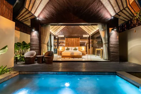 Luxurious tropical bedroom with king bed and open glass doors overlooking a lit private pool at night.