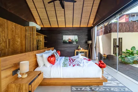 Romantic Balinese bedroom with wooden bed scattered in red rose petals, ceiling fan, and tropical garden view through sliding doors.