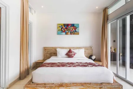 Modern tropical bedroom with wooden platform bed, white bedding, red accents, colorful abstract wall art, and beige curtains framing sliding glass doors.