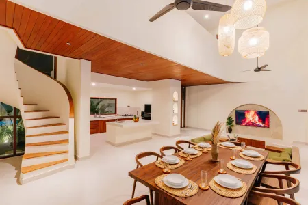 Modern tropical villa interior with open-plan living, dining, and kitchen, wooden table set for meal, curved staircase, ceiling fans, and lush greenery views. - Bali Villas