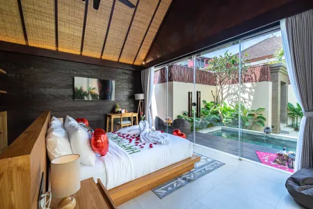 Romantic Bali villa bedroom with rose petal heart on white king bed, sliding doors to private plunge pool, and lush garden view.