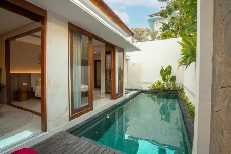 Serene private pool villa with open sliding doors to a bedroom, surrounded by tropical plants and stone walls under a blue sky.