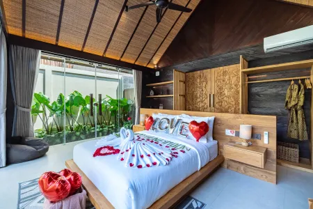 Romantic Balinese villa bedroom with king bed adorned in red rose petals, towel swans, "LOVE" pillows, and heart balloons, featuring wooden accents and tropical garden views.