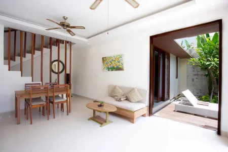 Modern tropical villa interior with wooden dining table, beige sofa, ceiling fans, and open doors to lush garden patio. - Bali Villas