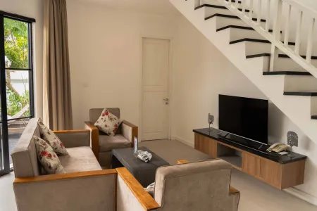 Modern living room with beige sofa, floral armchairs, coffee table, wall-mounted TV, and adjacent staircase with tropical garden view. - Bali Villas