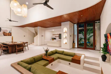 Spacious modern tropical villa interior with green L-shaped sofa, wooden dining table, open kitchen, and garden view through large doors. - Bali Villas