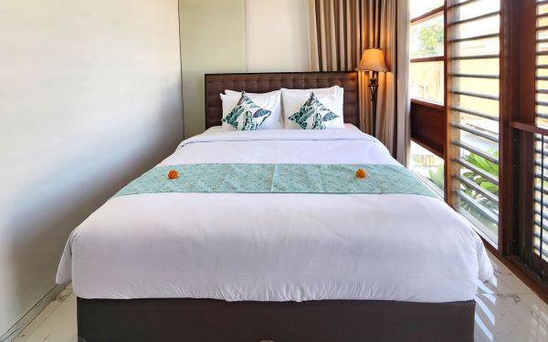 Elegant tropical bedroom with a white king bed, teal runner, and green leaf pillows, featuring large wooden louvered windows and soft lamp lighting.
