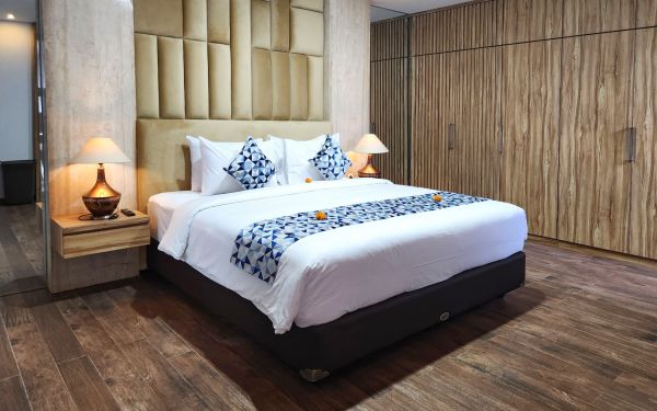 Elegant Balinese-style hotel bedroom with king bed, white linens, blue accents, frangipani petals, wooden walls, and bedside lamps.