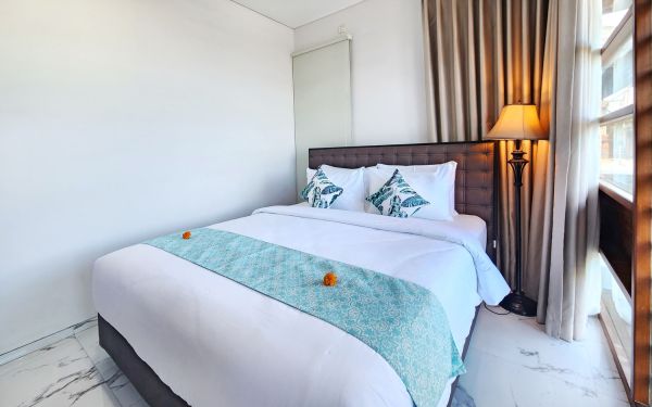 Cozy modern bedroom with a queen bed, teal runner adorned with frangipani flowers, leaf-patterned pillows, floor lamp, and sheer curtains in a tropical style.