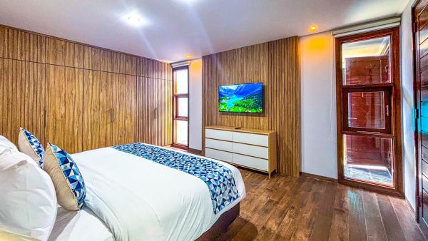 Modern bedroom with warm wooden walls and flooring, king bed with white sheets and blue duvet, white dresser, wall-mounted TV, and large wooden-framed windows.