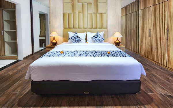 Elegant Balinese bedroom with king-sized bed, white linens, blue floral pillows, frangipani runner, bamboo wall panels, and warm lamp glow.
