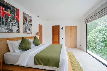 Spacious tropical bedroom with wooden bed, green throw pillows, vibrant wall art, and large windows overlooking lush greenery.