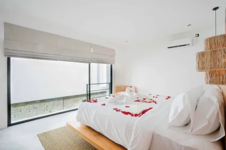 Romantic minimalist bedroom with white bed scattered in red rose petals, towel swan, large window view, and rattan lamps.