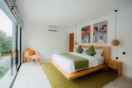 Modern tropical bedroom with platform bed in green linens, orange accent chair, abstract wall art, and large windows overlooking lush greenery. - Bali Villas