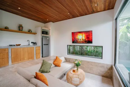 Modern tropical living room with wooden accents, cozy beige sofa with green and orange pillows, small kitchenette, and pool view through large windows. - Bali Villas