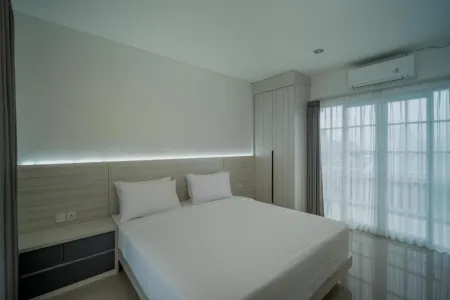 Modern bedroom interior featuring a white king-size bed with wooden headboard, LED lighting, bedside table, wardrobe, and sheer-curtained balcony doors.