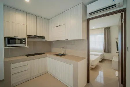 Modern white L-shaped kitchen with microwave, cooktop, and sink, opening to a cozy bedroom with twin beds in a studio apartment.