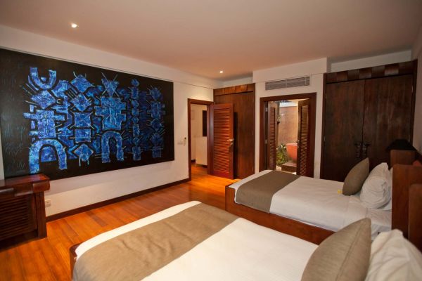 Spacious tropical bedroom with two neutral-bedded beds, large blue geometric wall art, and wooden doors and furniture.