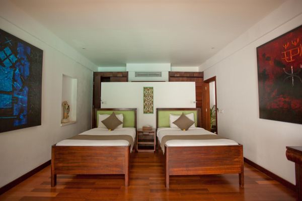 Spacious tropical twin bedroom with wooden beds, green cushions, abstract wall art, and wooden floors.