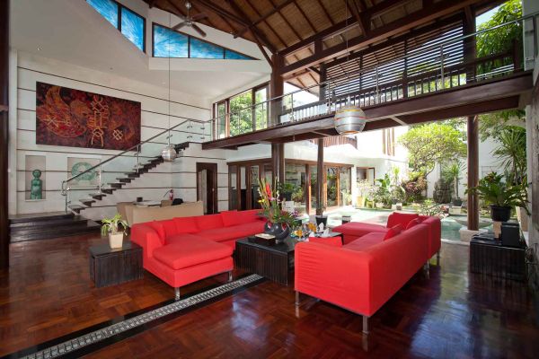Spacious open-plan living room in a modern Balinese villa with red L-shaped sofa, wooden beams, tropical plants, and garden views through glass walls. - Bali Villas