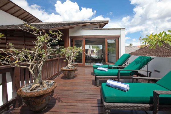 Luxurious Balinese villa rooftop terrace with green lounge chairs, white towels, potted frangipani trees, and sliding glass doors under a clear blue sky. - Bali Villas