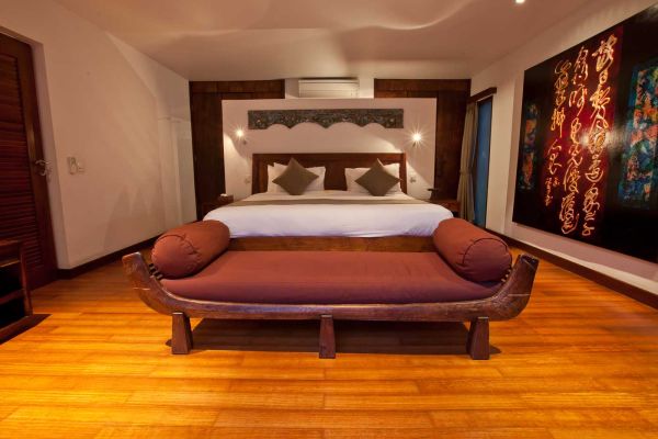 Luxurious Balinese-style bedroom with king bed, carved wooden benches, abstract wall art, and warm wooden floors.