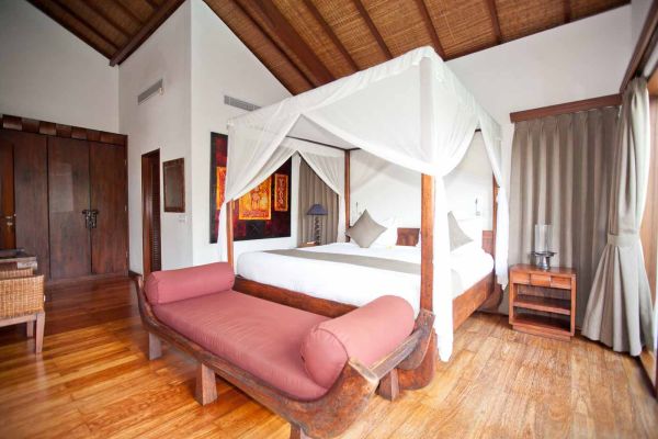 Luxurious Balinese bedroom interior with four-poster canopy bed, red chaise lounge, and wooden thatched ceiling.