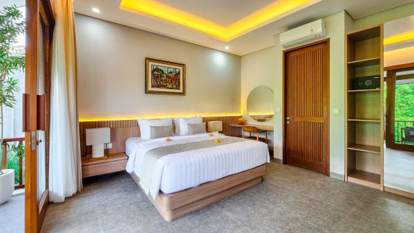 Elegant Balinese bedroom with wooden king bed, white linens, ambient yellow ceiling lights, and tropical garden view through sheer-curtained doors.