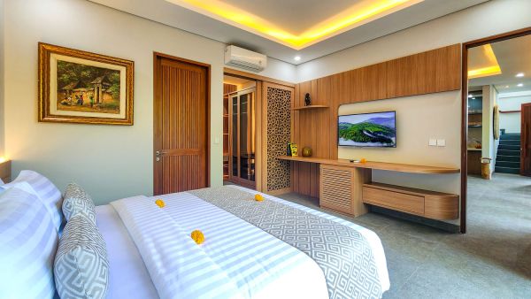 Spacious Bali villa bedroom with king bed adorned in frangipani flowers, wooden furnishings, wall-mounted TV, and balcony doors.