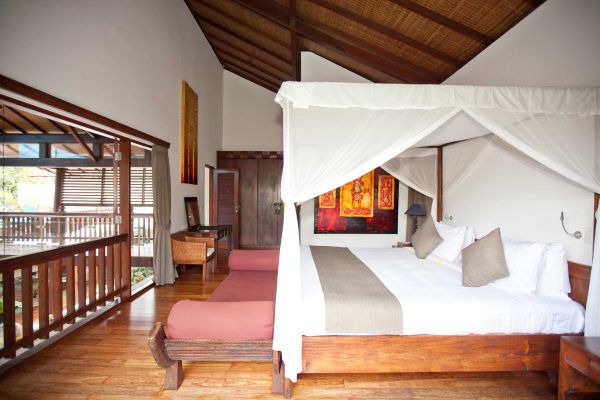 Luxurious Balinese villa bedroom featuring a white canopy four-poster bed, wooden floors, abstract wall art, and open balcony with tropical views.