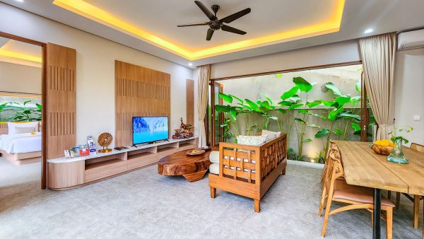Spacious Balinese villa living room with wooden sofa, TV, coffee table, dining set, and sliding doors to lush green garden. - Bali Villas